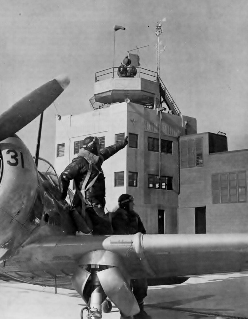A historic picture of two cadets sitting on a plane at the Bunker Hill NAS in 1943. The air control tower is seen in the background.