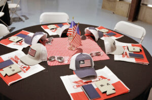 Picture of an empty table decorated with a flag-based centerpiece and at each seat a placemat, coozie, hat, and notepad.